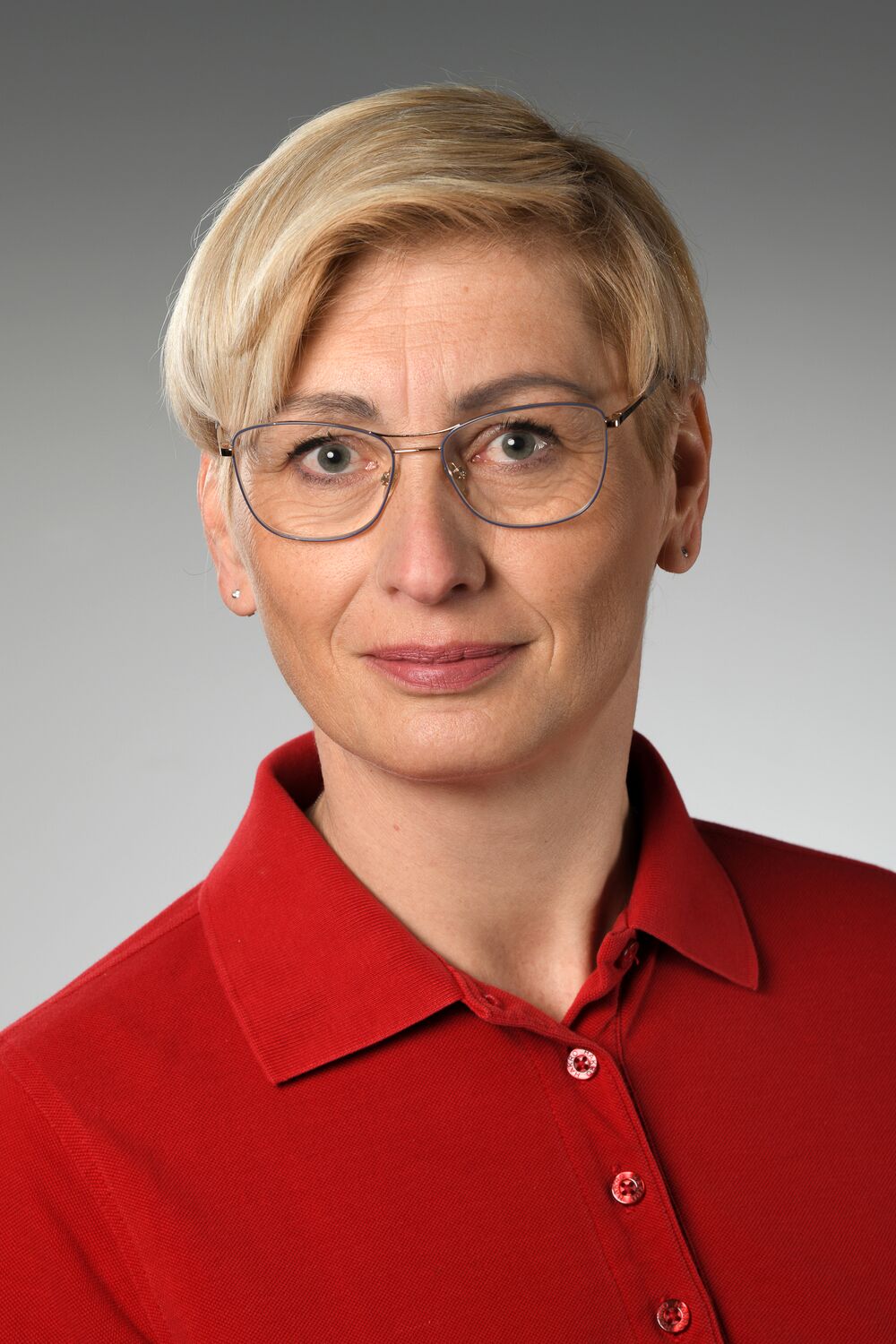 Portrait Frau Belz: Frau mittleren Alters mit kurzen, blonden Haaren und Brille. Sie trägt ein rotes Polo-Hemd und lächelt leicht in die Kamera.